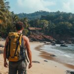 A traveler with a backpack walks along a scenic beach in Goa, India, with palm trees, golden sand, and a vibrant sunset in the background